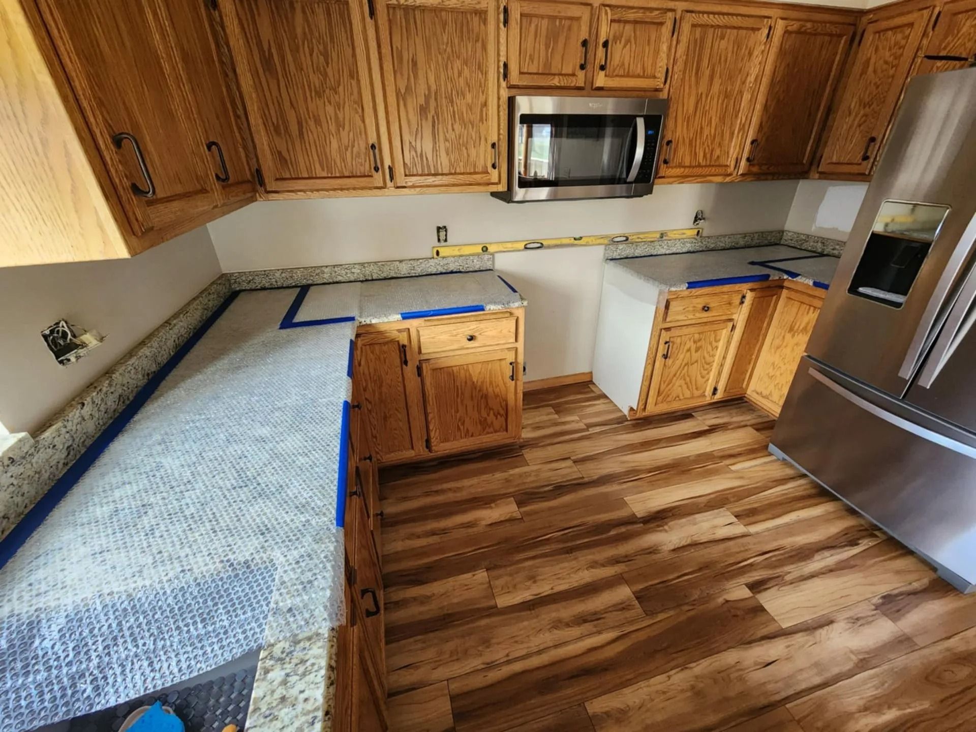 Small kitchen with wood cabinets, stainless refrigerator, microwave, and blue-taped countertops under renovation.