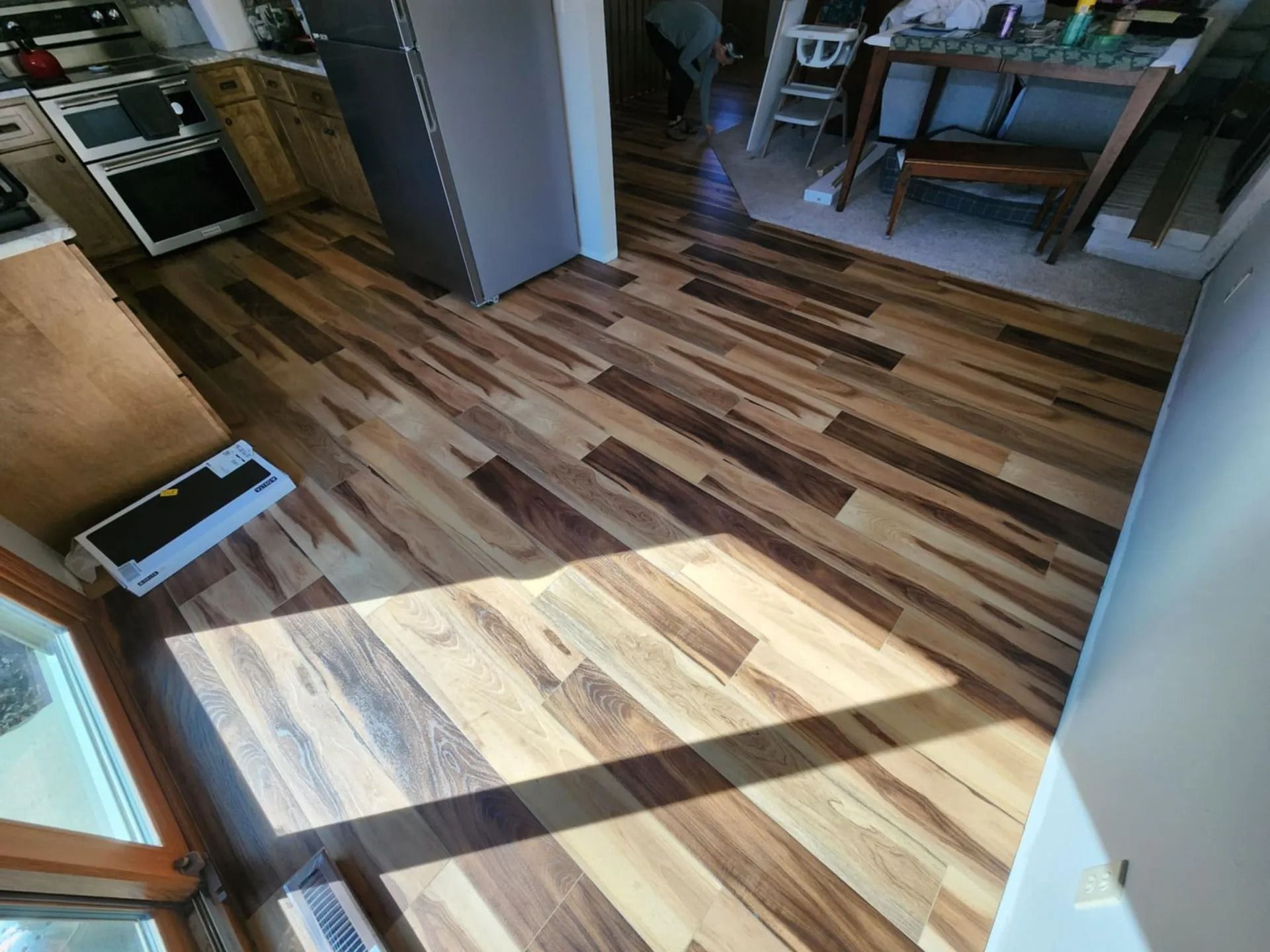 Kitchen and dining area with wood-patterned floor, sunlit doorway, and dining table in the background