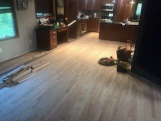 Empty kitchen with light wood flooring, scattered items, and dark cabinets near a window.