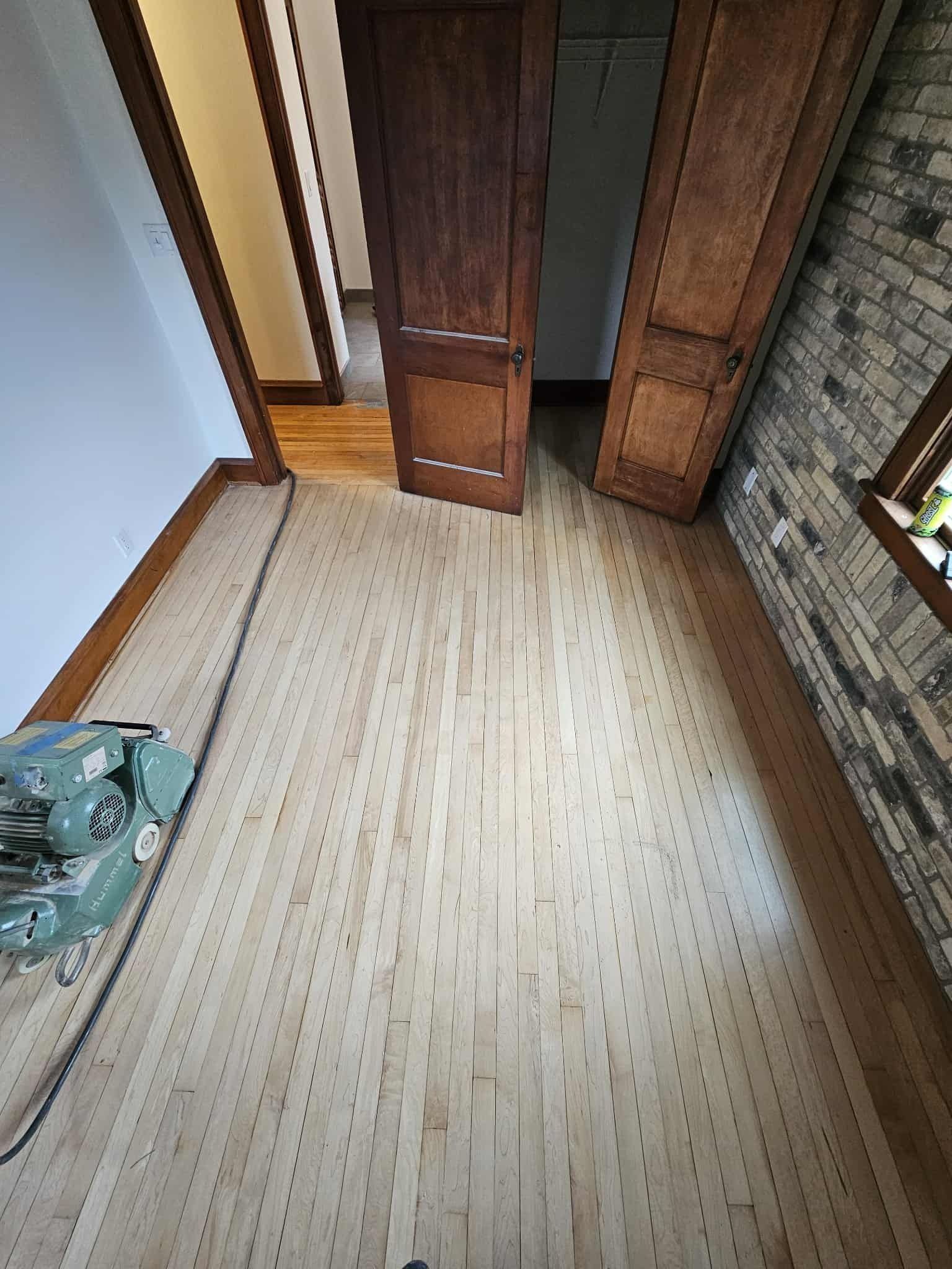 Empty room with light wood floor, open wooden doors, and a brick wall with a window.