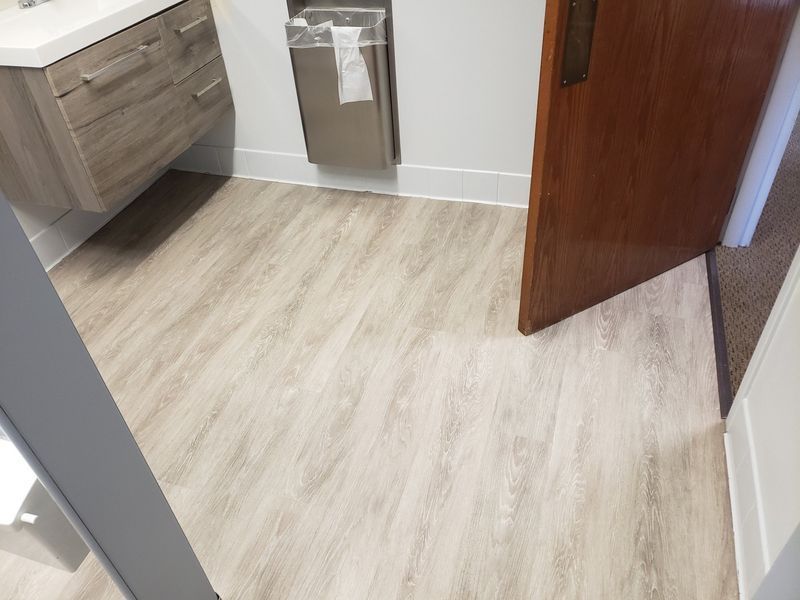 Small bathroom with light wood flooring, vanity, trash can, and an open wooden door.