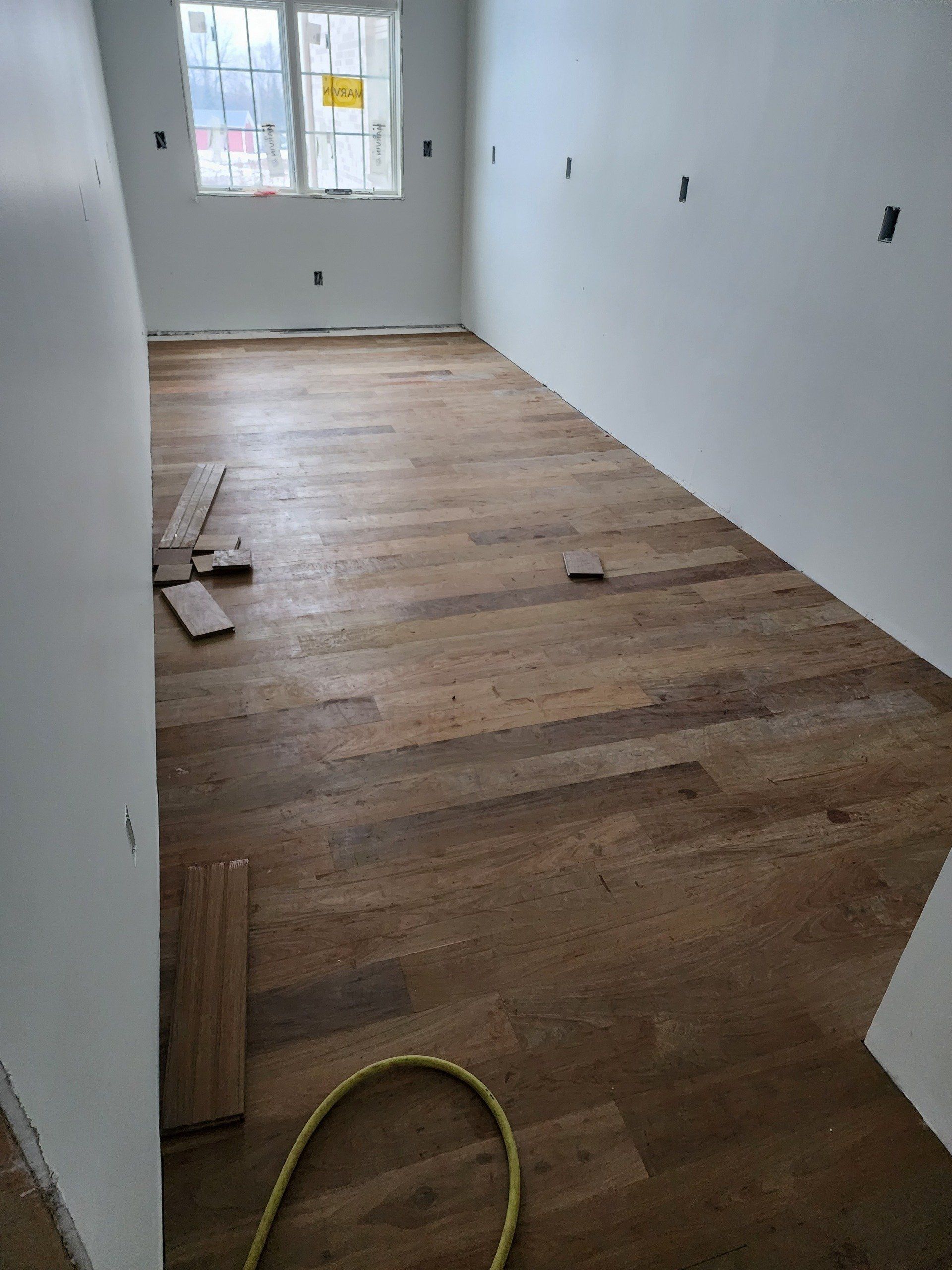 Empty room with unfinished wooden floor, white walls, and a window at the far end.