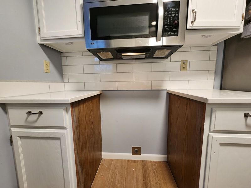 Empty kitchen nook with white cabinets, stainless microwave, and wood-look floor panels under the counter area