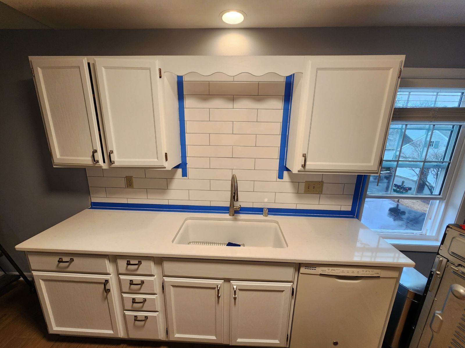 White kitchen cabinets and counters with blue painter’s tape around a sink and window backsplash under renovation