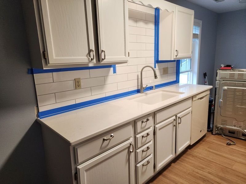 Partially remodeled kitchen with white cabinets, gray walls, and blue painter’s tape marking surfaces