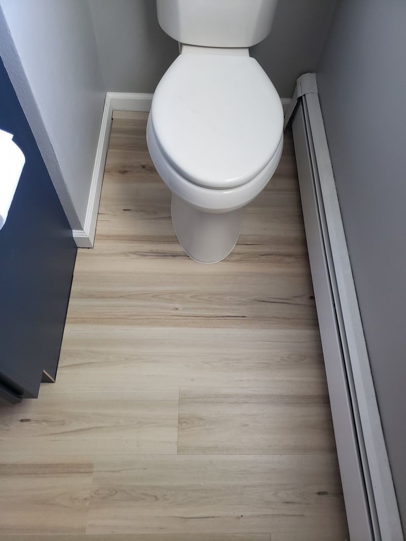 Small bathroom with a toilet on light wood flooring between gray walls.