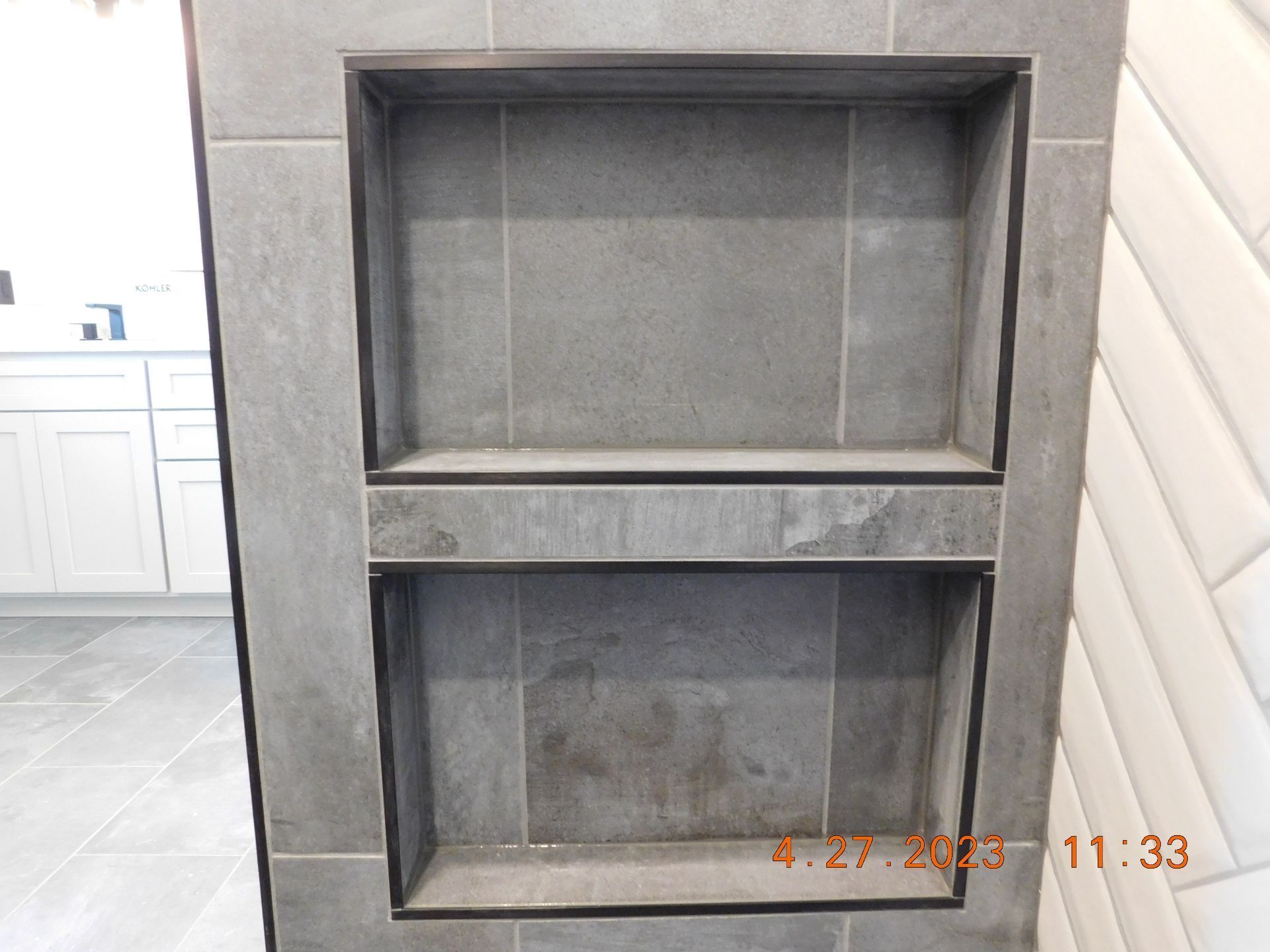 Gray built-in wall niche with two open shelves and a lower storage compartment in a tiled room.