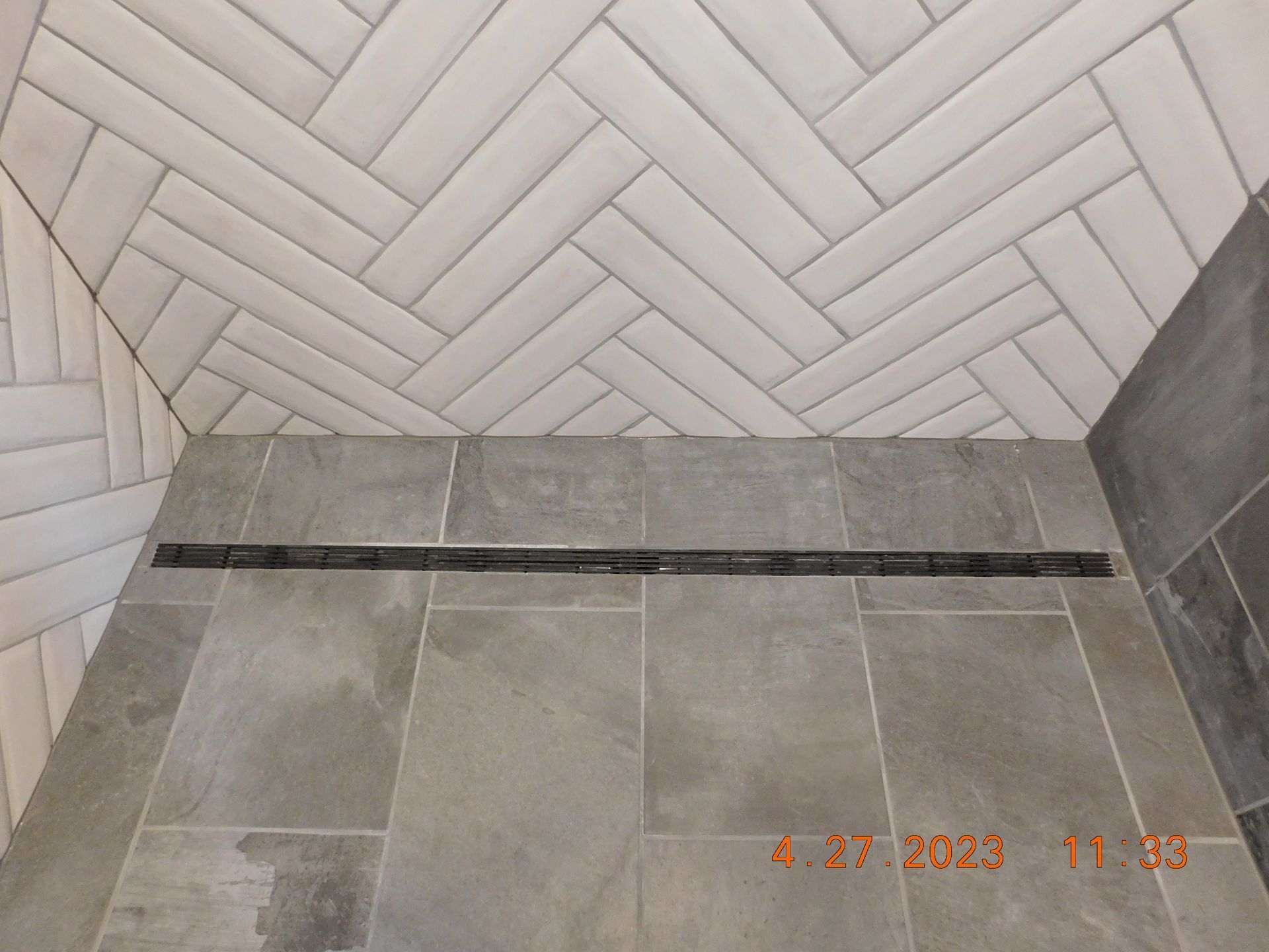 Gray tiled shower floor and herringbone white wall tiles with a dark grout drain strip nearby