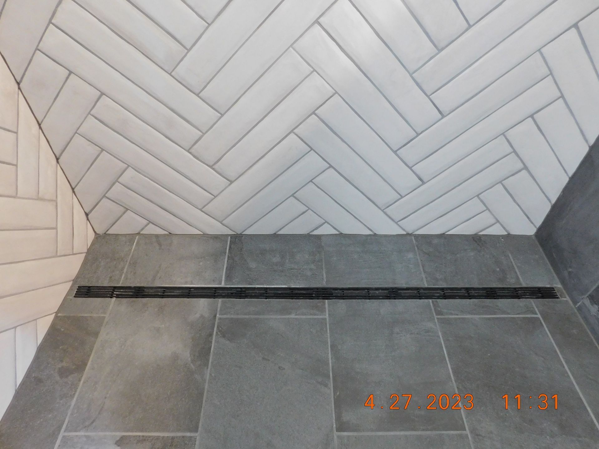Gray tiled shower floor and herringbone white wall tiles with a dark bench ledge.