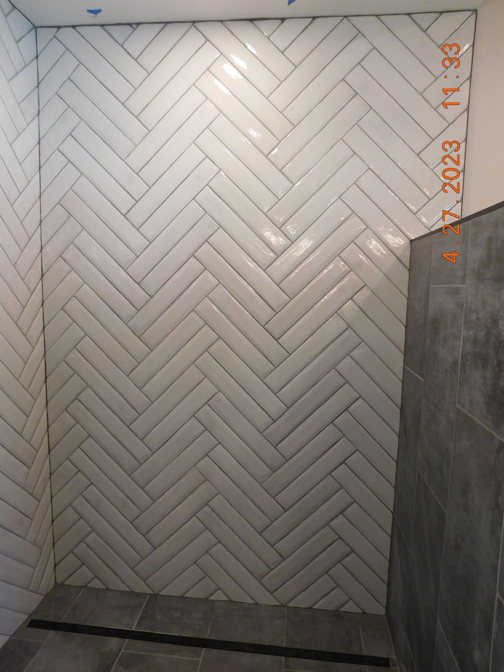 White herringbone tile wall in a shower alcove with gray side panels and ceiling lights.