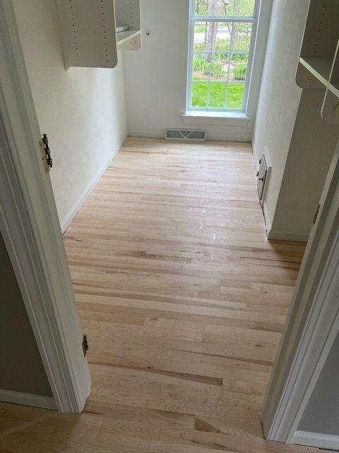 Empty narrow room with light wood floors, white walls, and a window at the far end.