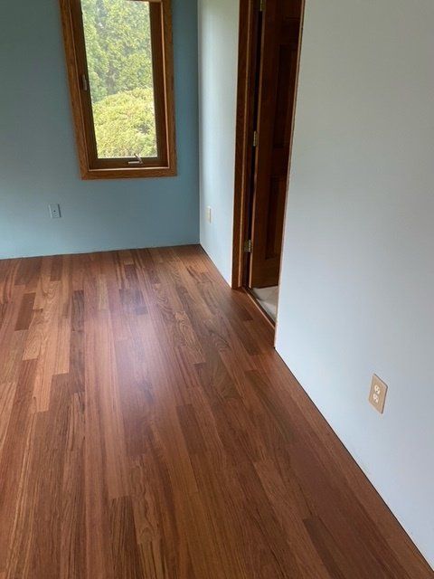 Empty room with wood floor, white walls, and a window looking out onto greenery.