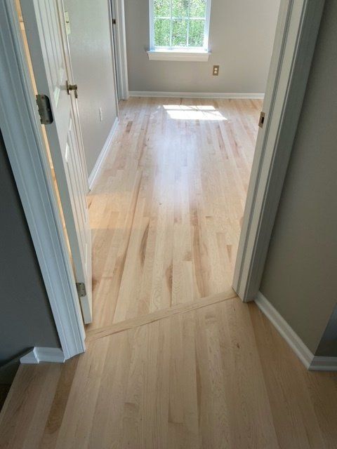 Empty hallway with light wood flooring leading to a bright room with a window.