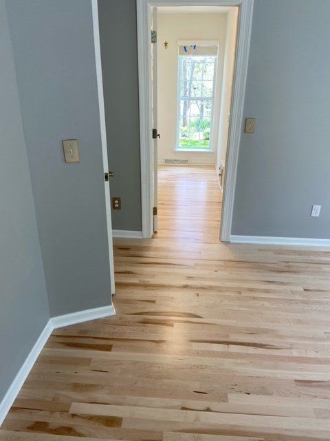 Empty hallway with light wood floors, gray walls, and a doorway looking into a bright room beyond