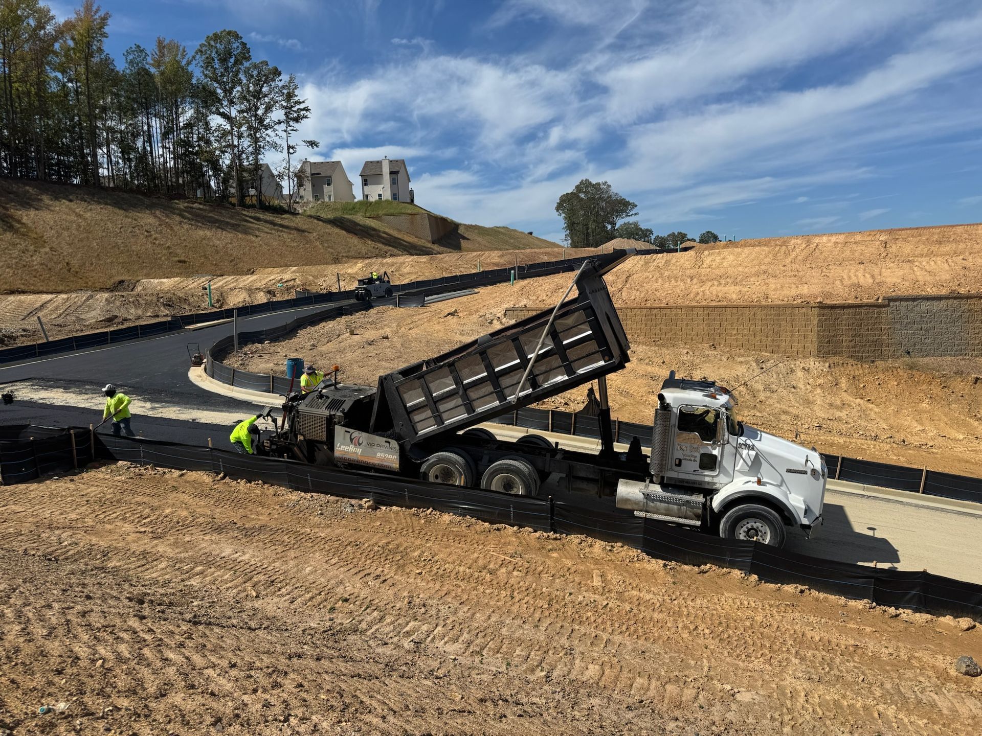 Asphalt being poured by a dump truck on a new road under construction. Workers in safety vests are present.