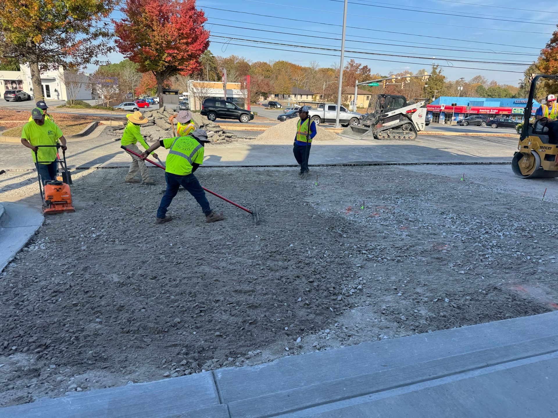 Construction workers laying gravel in a parking lot; some using hand tools, one operating a compactor.