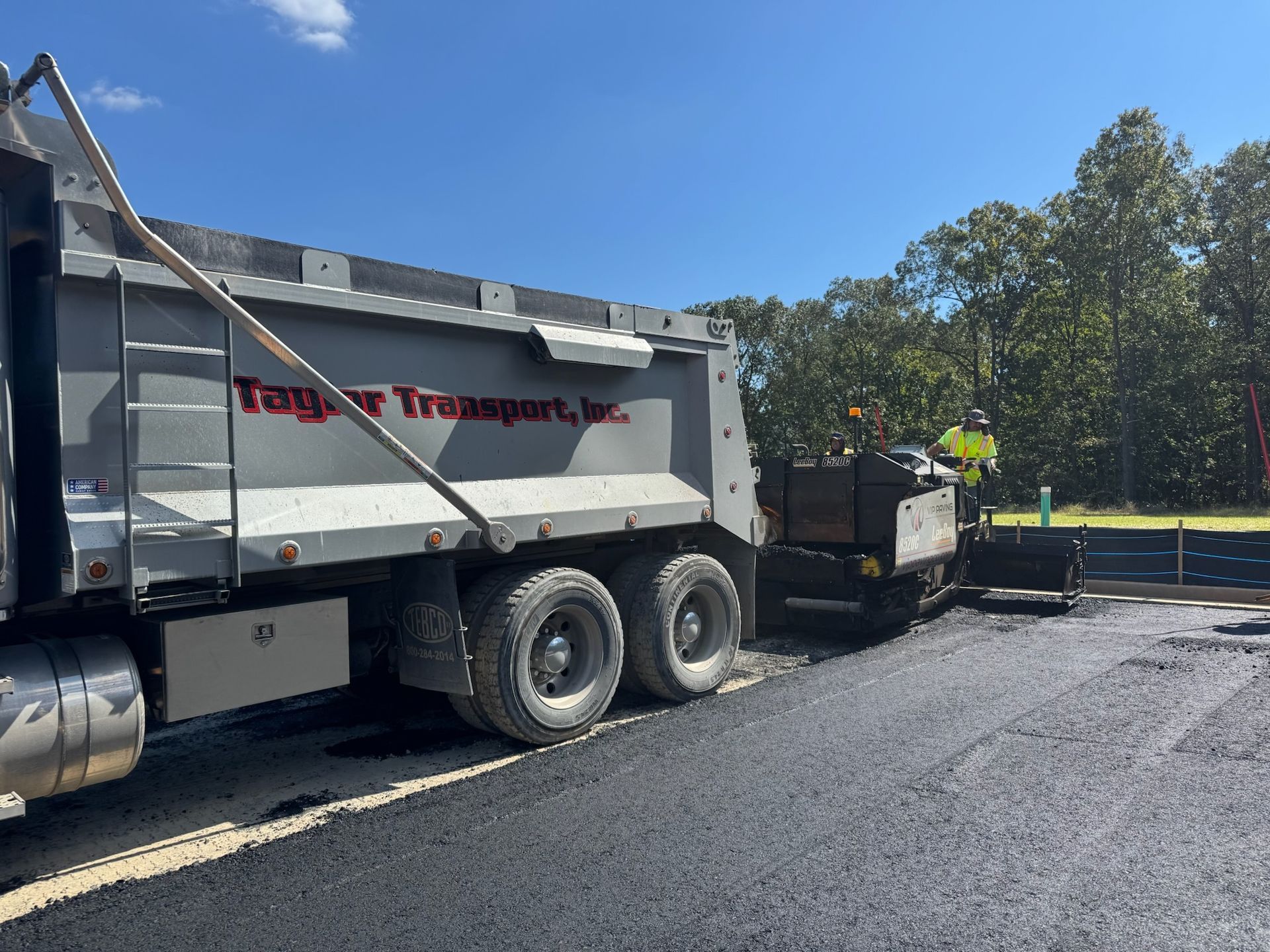 Dump truck unloading asphalt onto a paving machine on a sunny day.