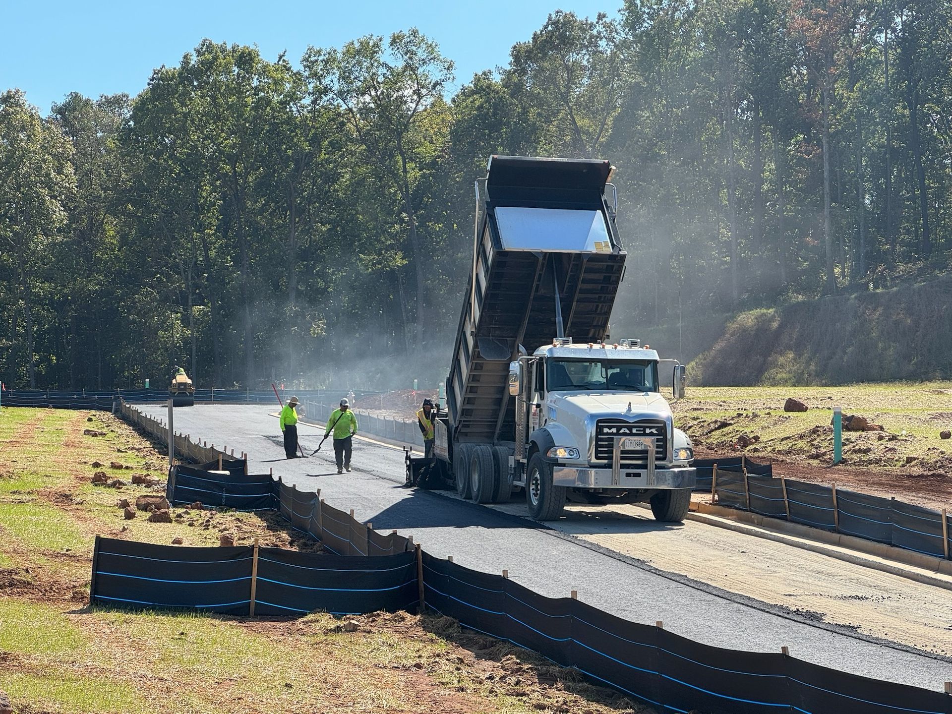 Dump truck unloading gravel on a road under construction; workers use shovels to level it. Forest in background.