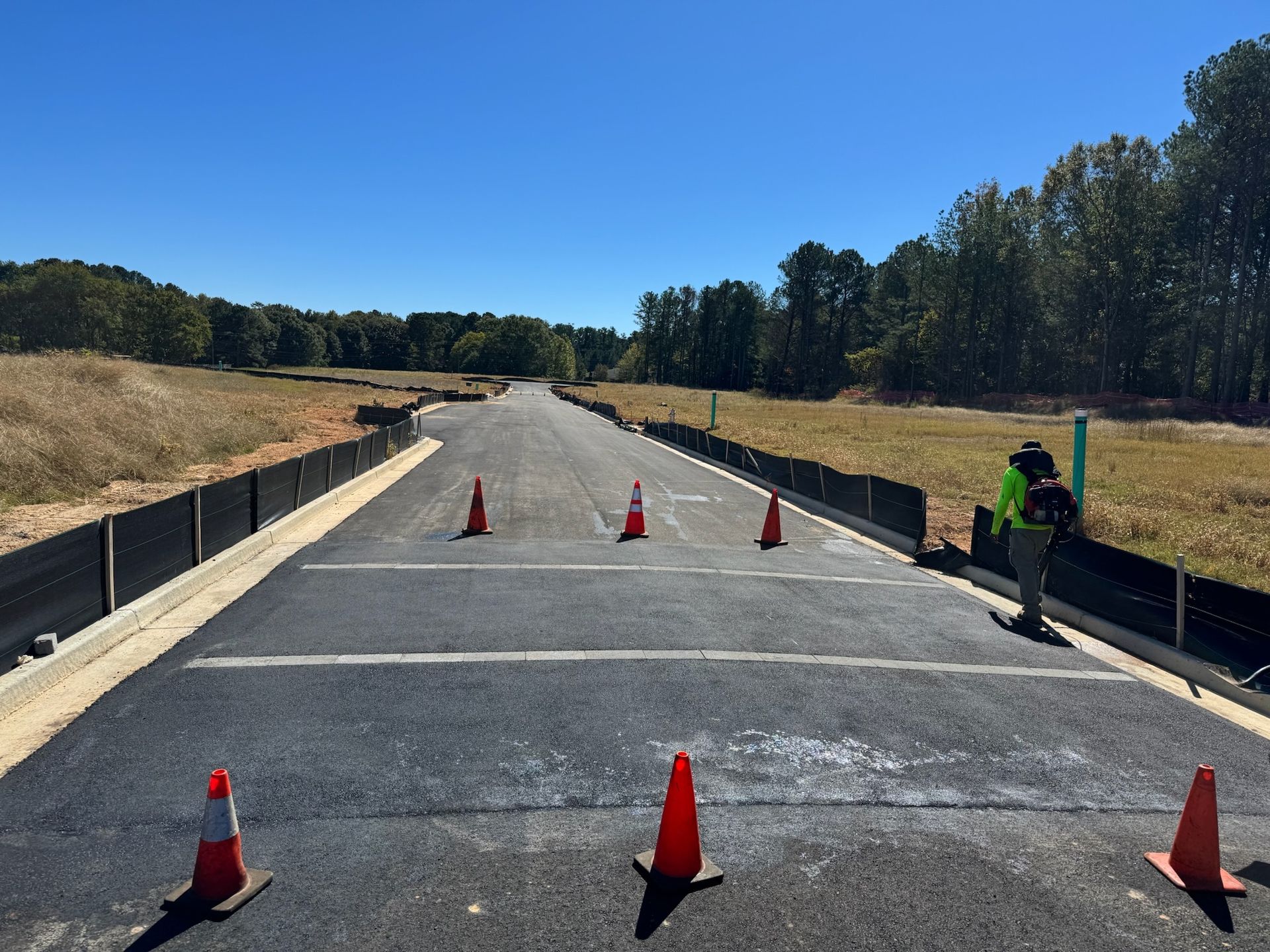 Newly paved road with orange cones and construction fencing, person on right, field and trees in the background.