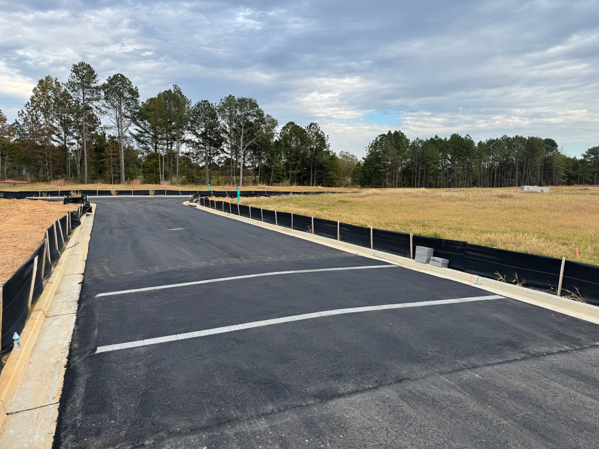 Paved road in a construction area with a grassy field and trees in the background under a cloudy sky.
