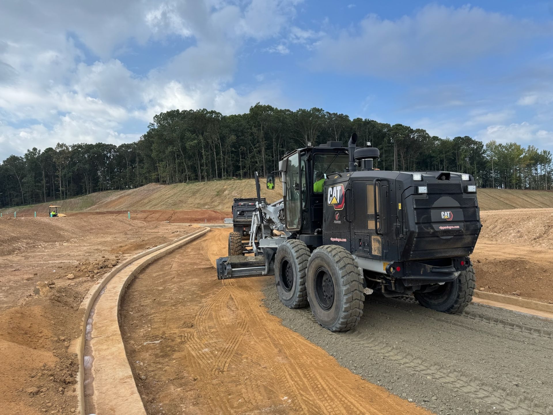 A motor grader compacts gravel on a construction site next to a curved ditch under a cloudy sky.