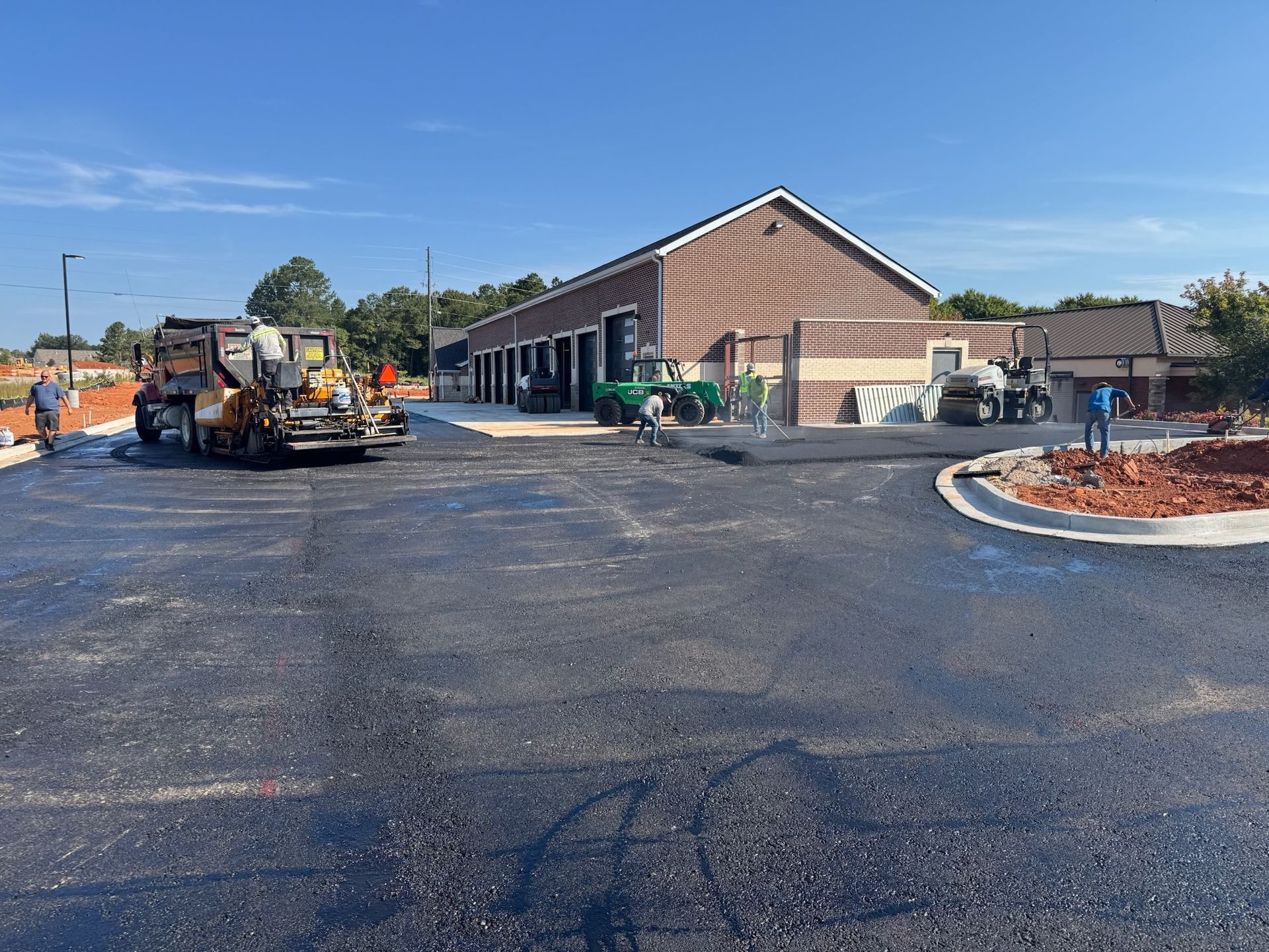 Asphalt paving a parking lot in front of a building; construction workers and machinery are present.