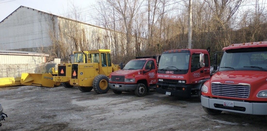 A yellow construction tractor parked next to three red trucks in a gravel lot with trees and a building in the background.