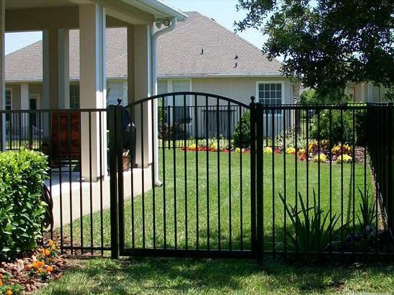 Ornamental fence and gate enclose a grassy backyard, with a house visible in the background.