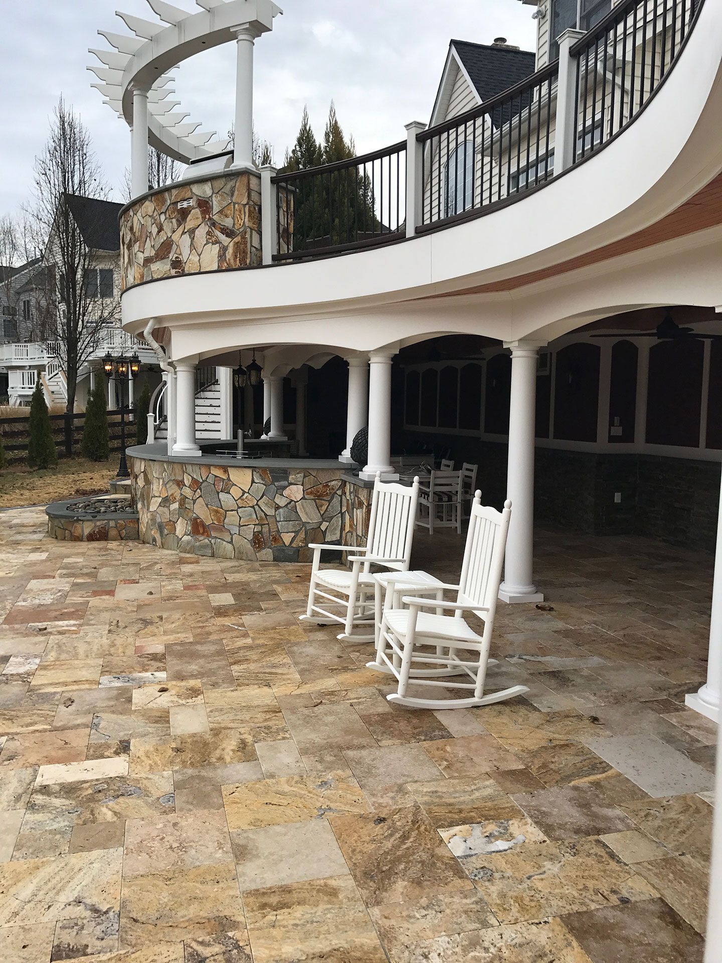 Two white rocking chairs on a stone patio beneath a covered outdoor space with pillars. A stone wall and a curved balcony are in the background.