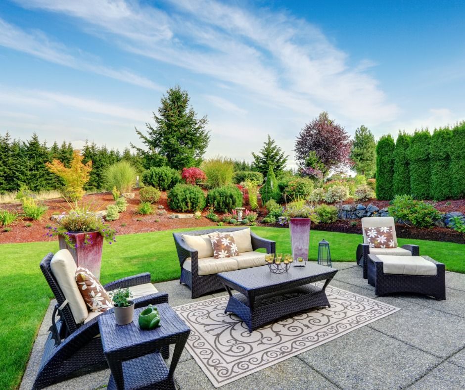 Patio with dark wicker furniture, beige cushions, and patterned rug on a stone surface, surrounded by lush green landscaping under a blue sky.