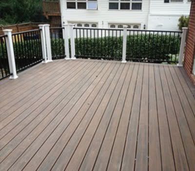 Trex deck with brown planks and a black railing with white posts, beside a house with a green hedge.