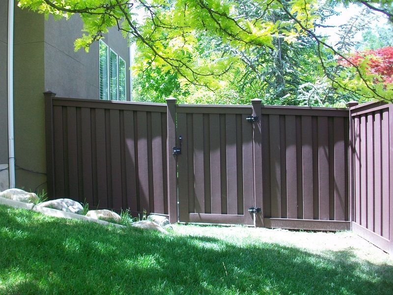 Brown trex fence with gate in a grassy yard, next to a building and trees.