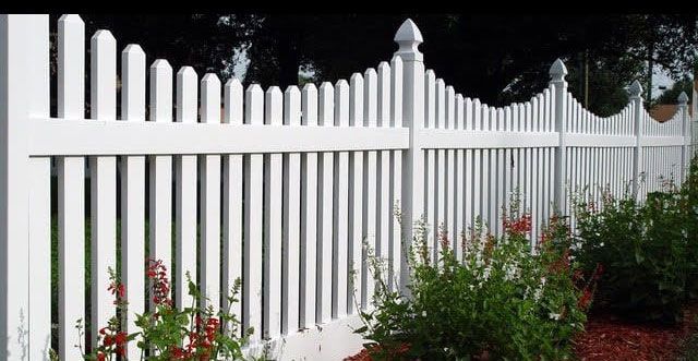 White vinyl fence with decorative posts in front of green and red plants, with trees in the background.