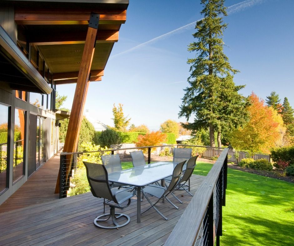Outdoor wooden deck with glass table and chairs overlooking a green lawn with trees in the background under a blue sky.