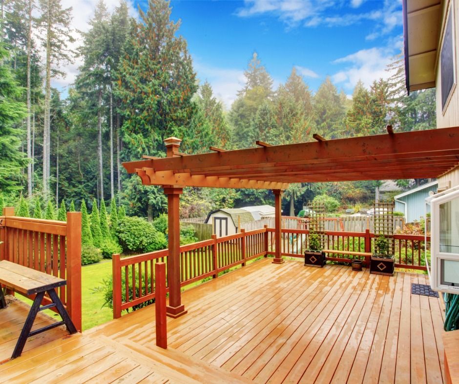Wooden deck with pergola, overlooking a green yard and trees. A bench sits on the deck's edge.