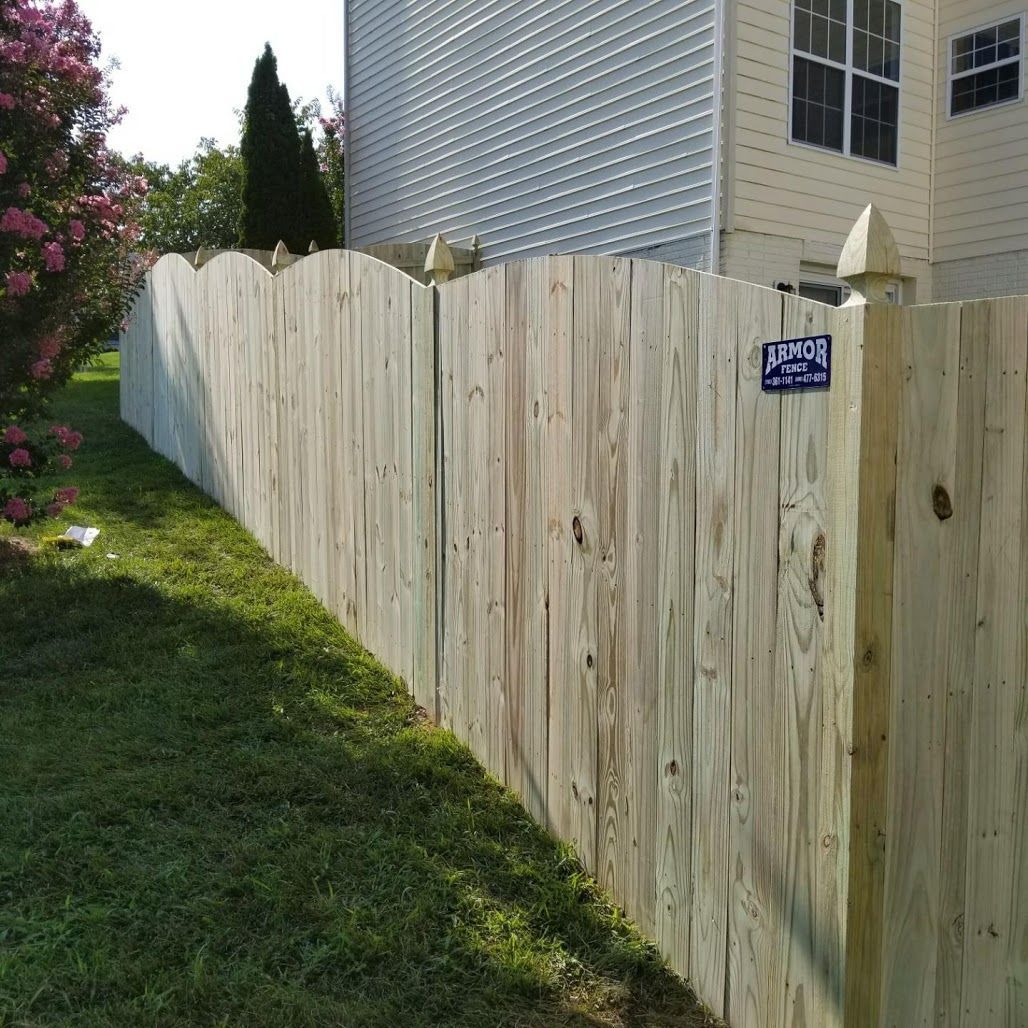 A wooden fence with a scalloped top separates a grassy lawn from a house. The fence is made of light-colored vertical boards.