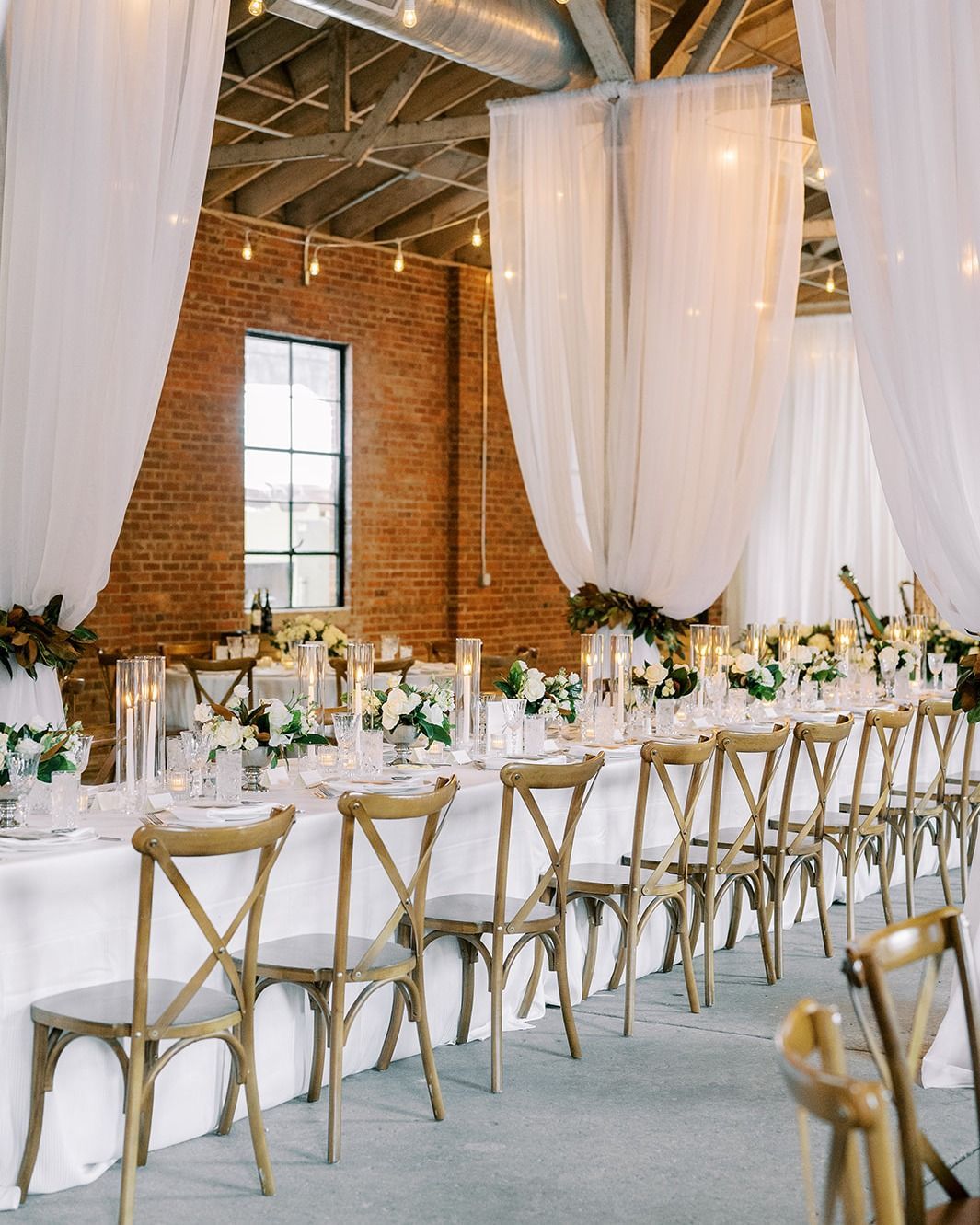 Wedding reception setup: long table with white linens, cross-back chairs, and floral centerpieces; draped fabric and brick wall.