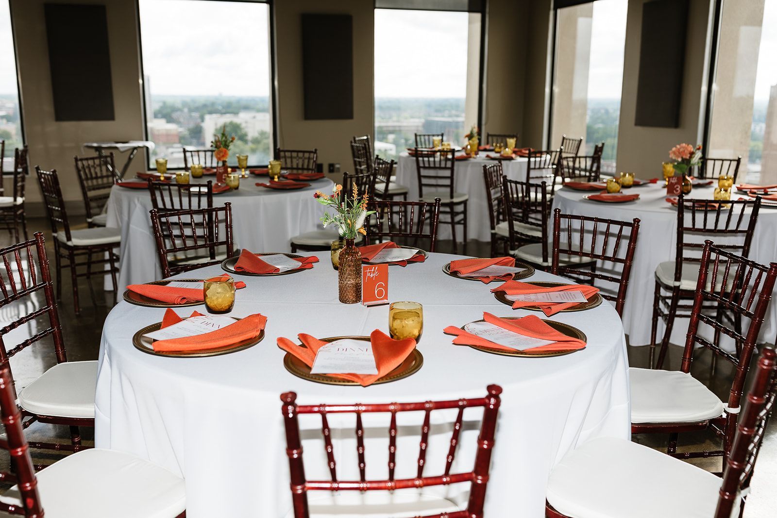 Round tables set for a party in a room with a city view. Tables have white tablecloths, orange accents, and brown chairs.