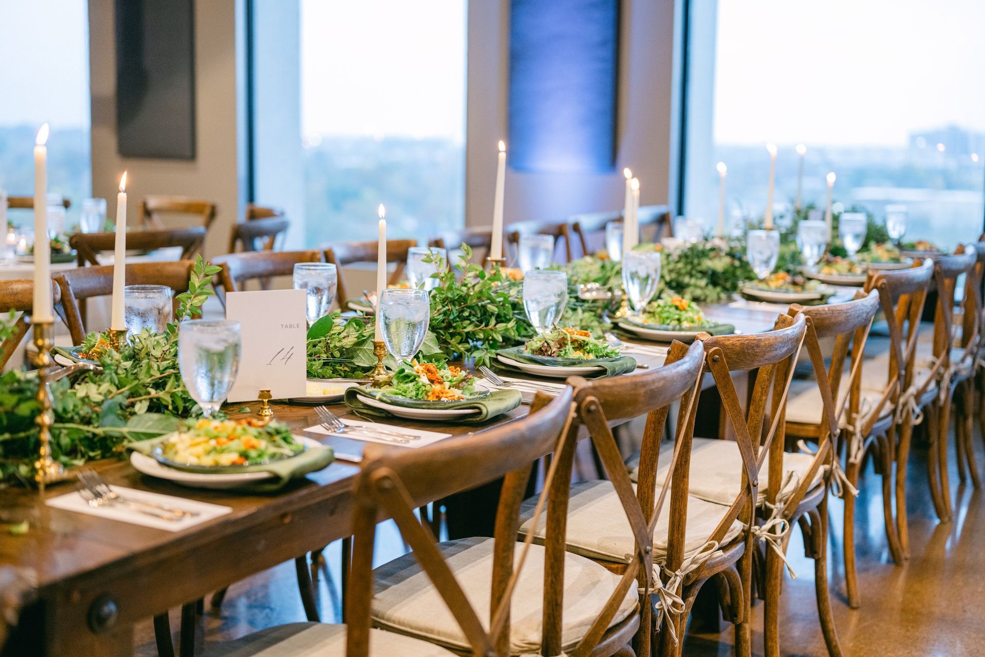 Long wooden banquet table set for a formal event, with greenery, candles, and views out of windows.