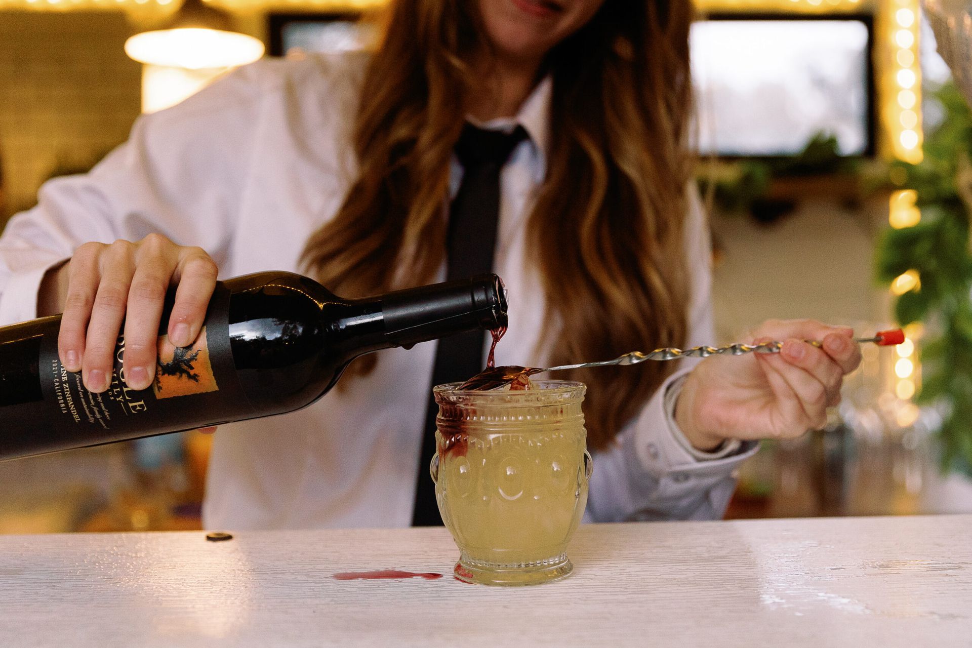 Bartender pouring red wine into a cocktail glass, mixing the drink with a long spoon. She wears a white shirt and tie.