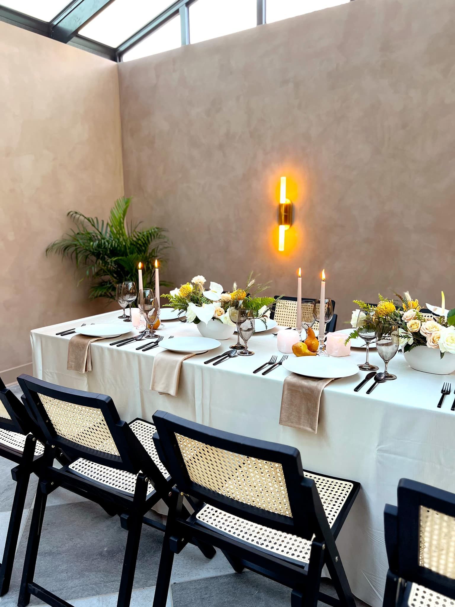 A well-set outdoor dining table with candles, flowers, and black chairs. Lit by a wall-mounted fixture with a neutral wall backdrop.