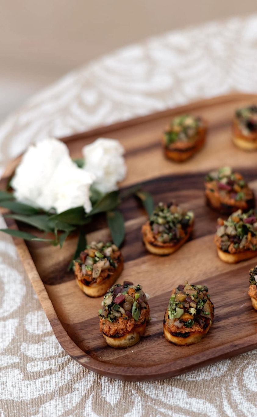 Wooden tray with appetizers topped with pistachios, near a white flower.