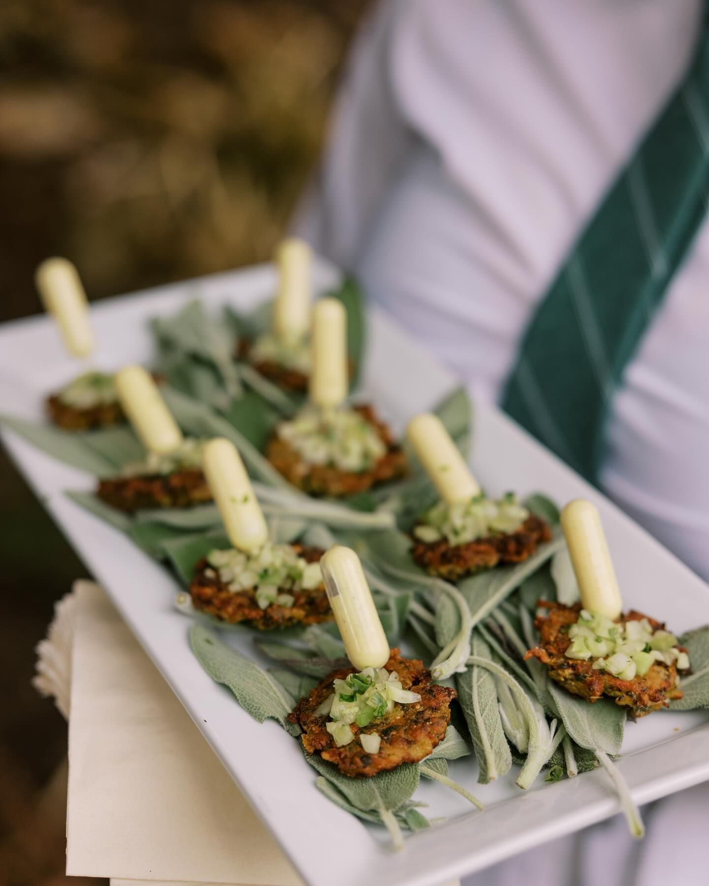 A person holding a platter of appetizers: fried cakes topped with green relish and a cylindrical garnish on sage leaves.