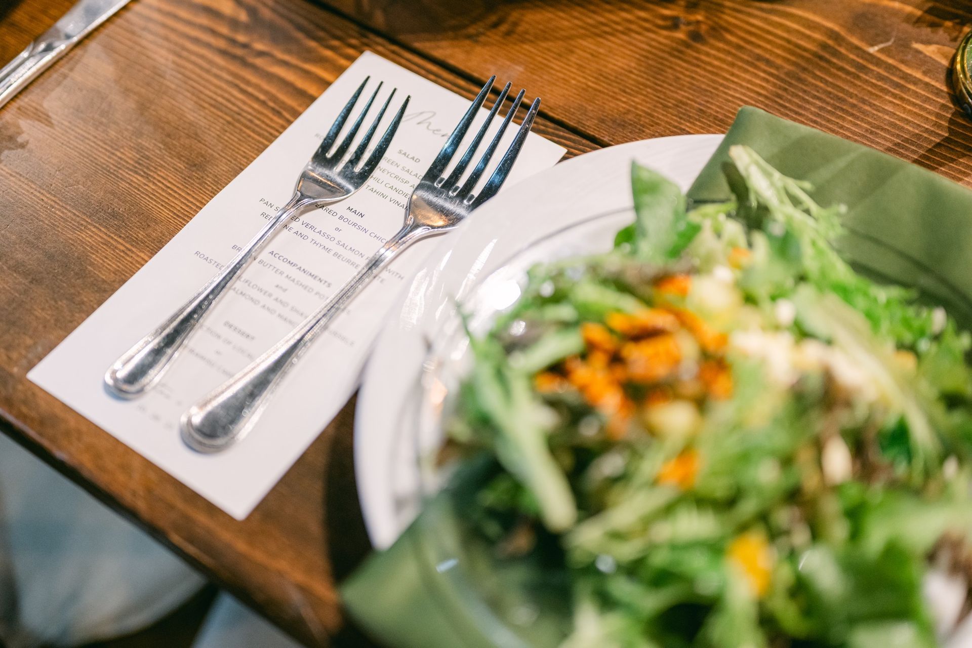 Two forks rest on a wooden table next to a plate of salad with a green napkin and menu.