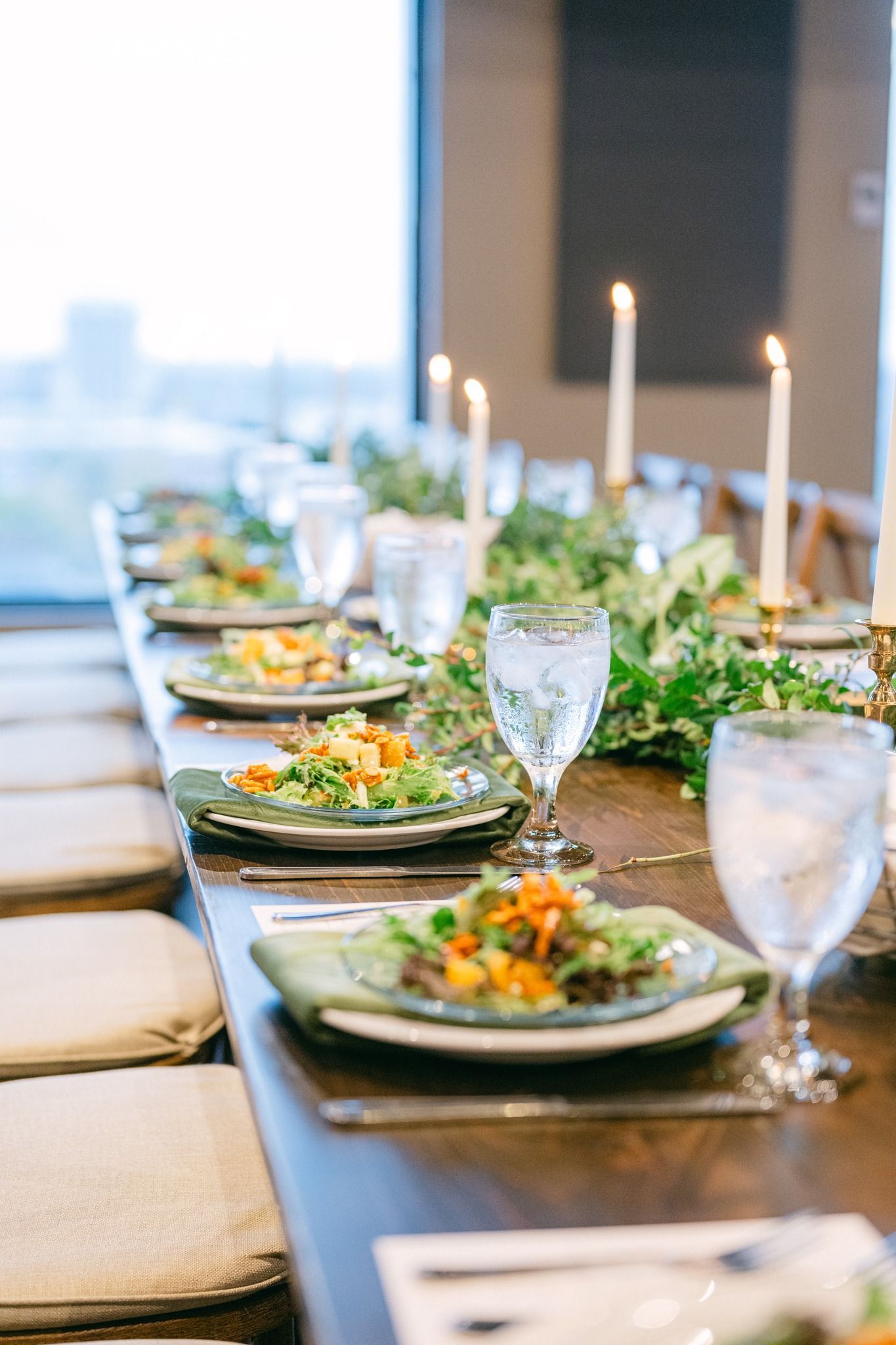 Long wooden table set for a meal, with plates of food, glasses of water, and lit candles. Greenery runs down the center.