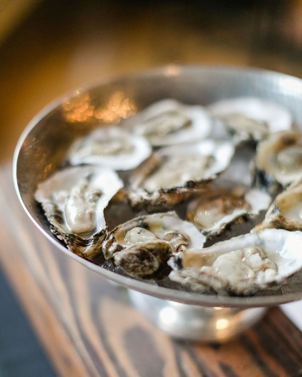 A silver platter of fresh oysters on ice, set on a wooden table.