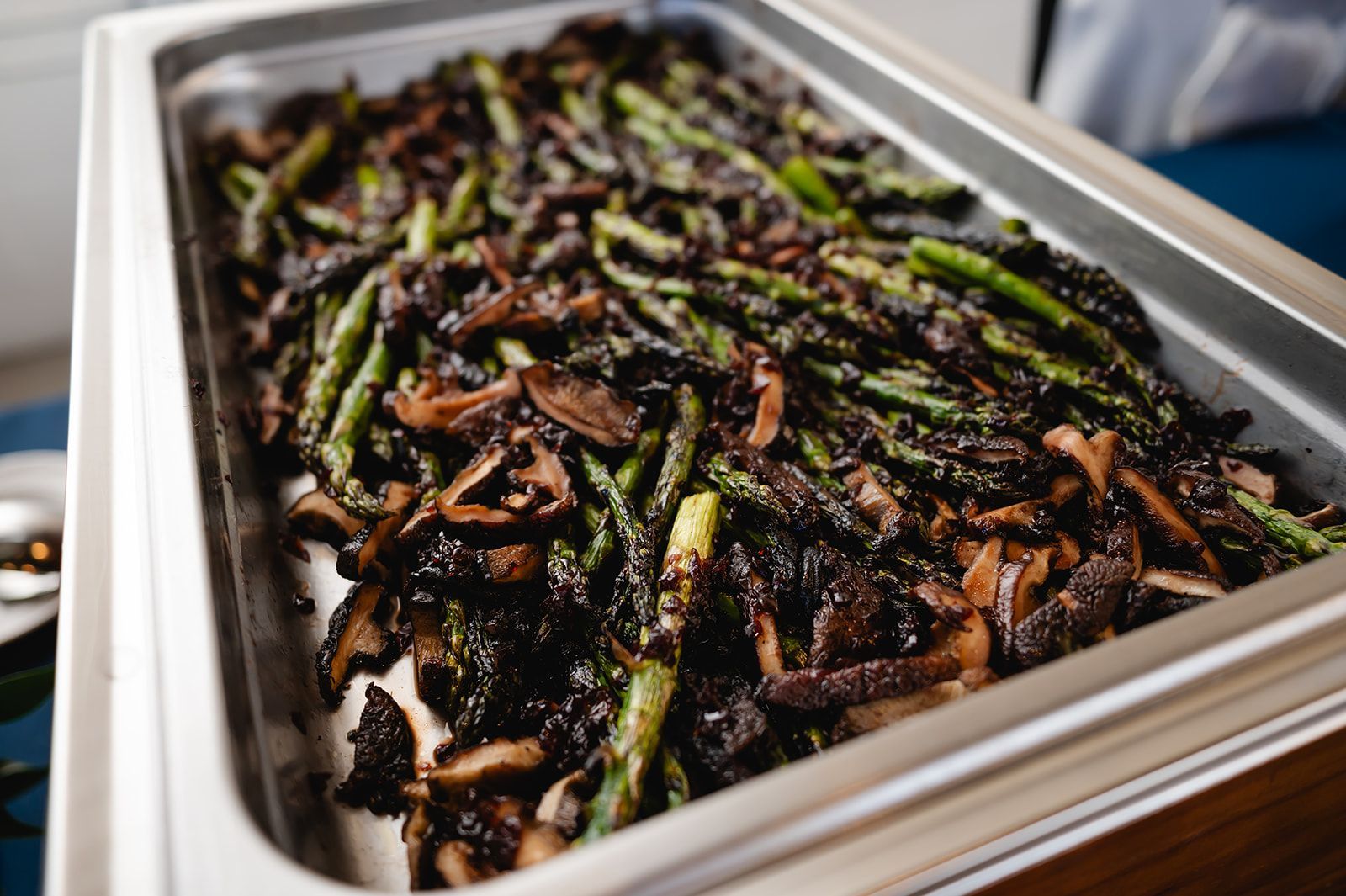 A stainless steel chafing dish filled with roasted asparagus and mushrooms. The dish is on a table.