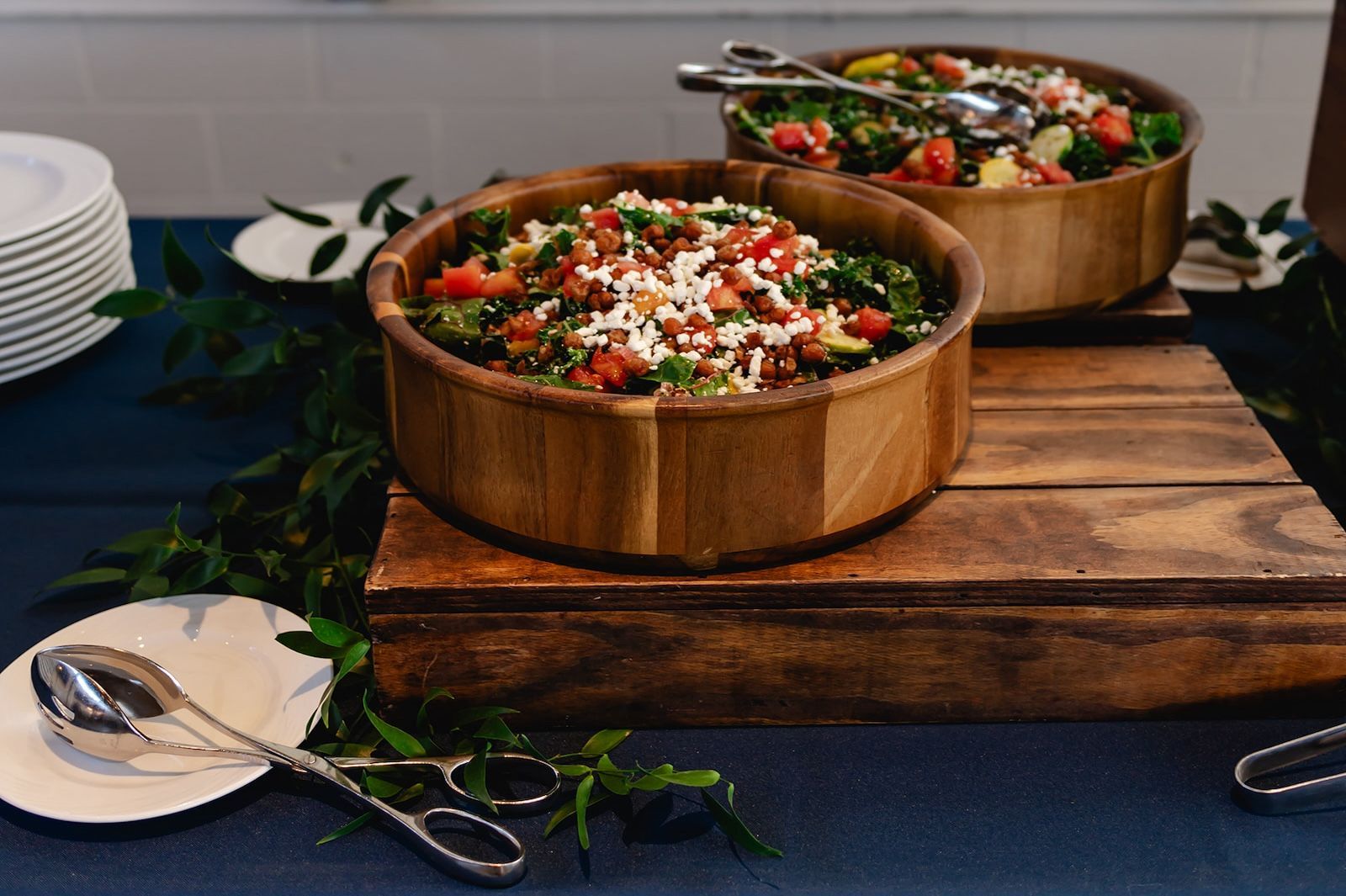 Two wooden bowls of salad topped with cheese and tomatoes on a wooden platform, with serving utensils, plates, and greenery on a blue tablecloth.
