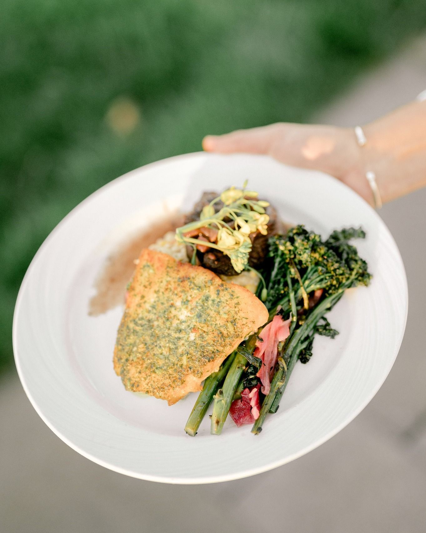 A person holding a white plate with a meal, including salmon, greens, and other vegetables.