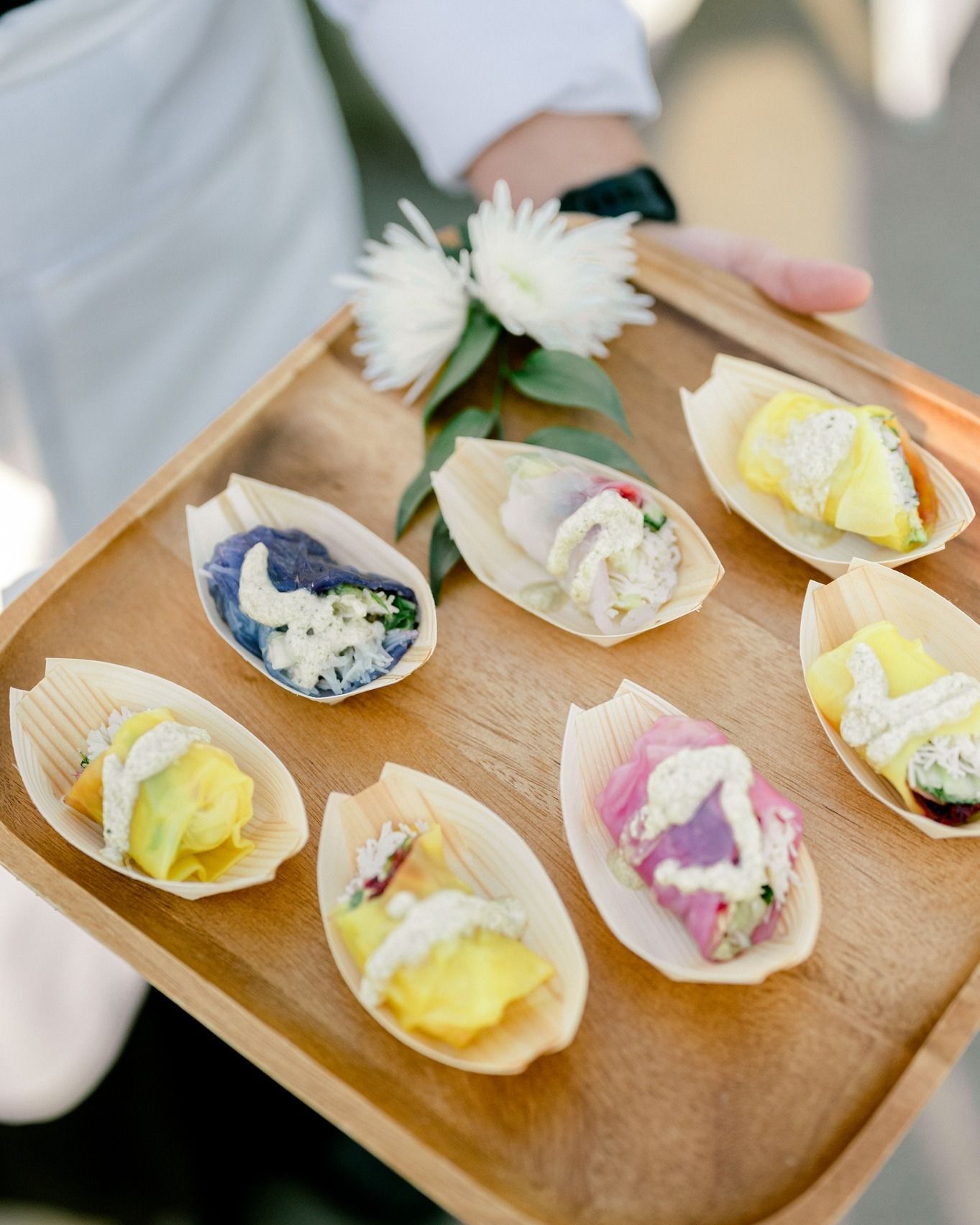 A wooden tray holds several colorful, bite-sized appetizers in leaf-shaped dishes. A server's arm and flowers are partially visible.